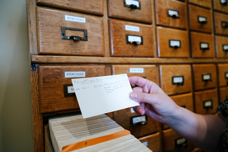 Person using a library catalog card from a wooden drawer system to retrieve bibliographic information.