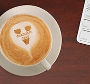 A cup of coffee with latte art displaying the "Graduate Café" logo, placed on a wooden table next to a white computer keyboard.