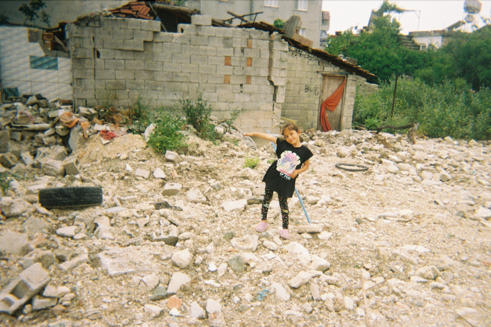 A person stands amidst rubble and debris in front of a partially demolished brick building. The area is scattered with rocks and broken bricks. Vegetation and other buildings are visible in the background.