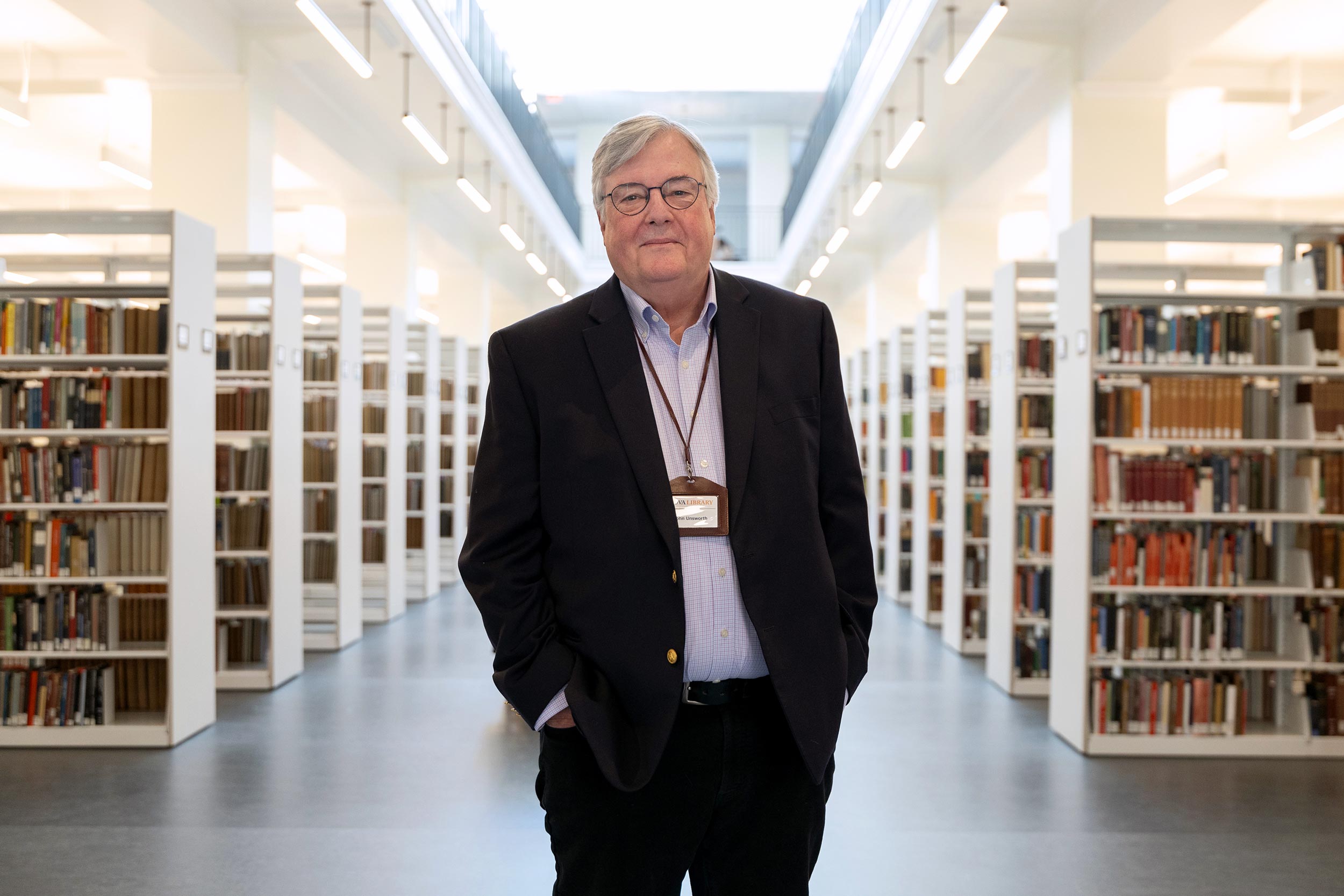 A man in a suit stands in the center of a large airy space, filled with book shelves. 