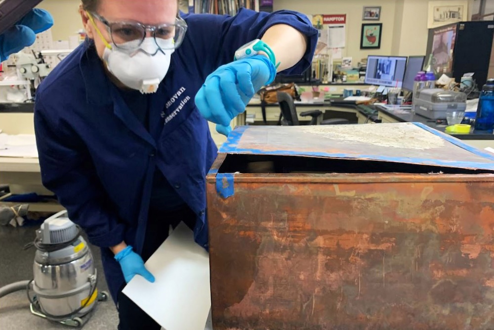A person wearing protective gear examines an open metal time capsule in a cluttered room. The individual is holding a document and is equipped with safety glasses, gloves, and a mask. Shelves, a vacuum cleaner, and a computer are visible in the background.