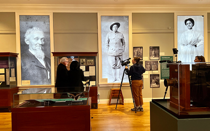 A view of the exhibition room showing large portraits behind John Edwin Mason, who is being interviewed in front of a camera