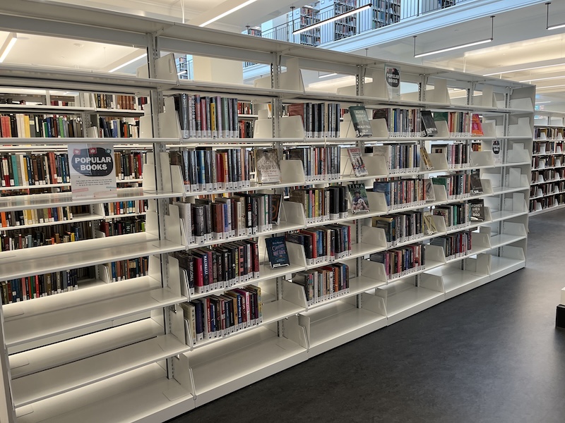 Interior of a modern library showing rows of bookshelves filled with books, labeled "Popular Books" in one section. Brightly lit environment with visible walkways among the shelves.