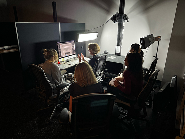 Four individuals working around a computer in a dimly lit room, discussing a project displayed on the screen. In the background is a large scanner.