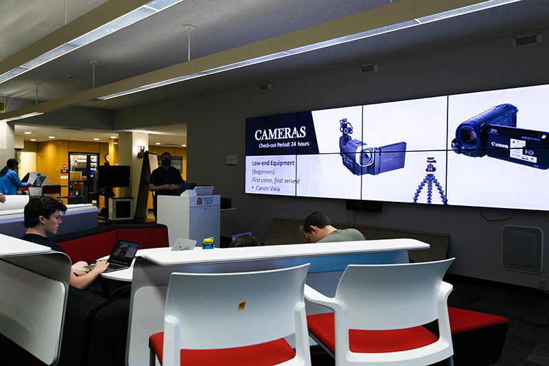 Interior of a library teaching space with people using laptops at desk pods. Two large screens display advertisements for cameras, specifically Canon models, with information on equipment loans.