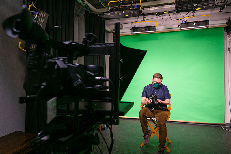 A person sits on a stool in a studio with a green screen background, holding a camera, with professional lighting equipment and a video camera in the foreground.