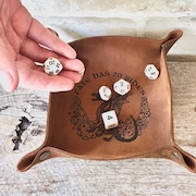 A hand holding dice over a leather rolling tray embossed with a decorative dragon design and the text "D&D The 20-Sided."