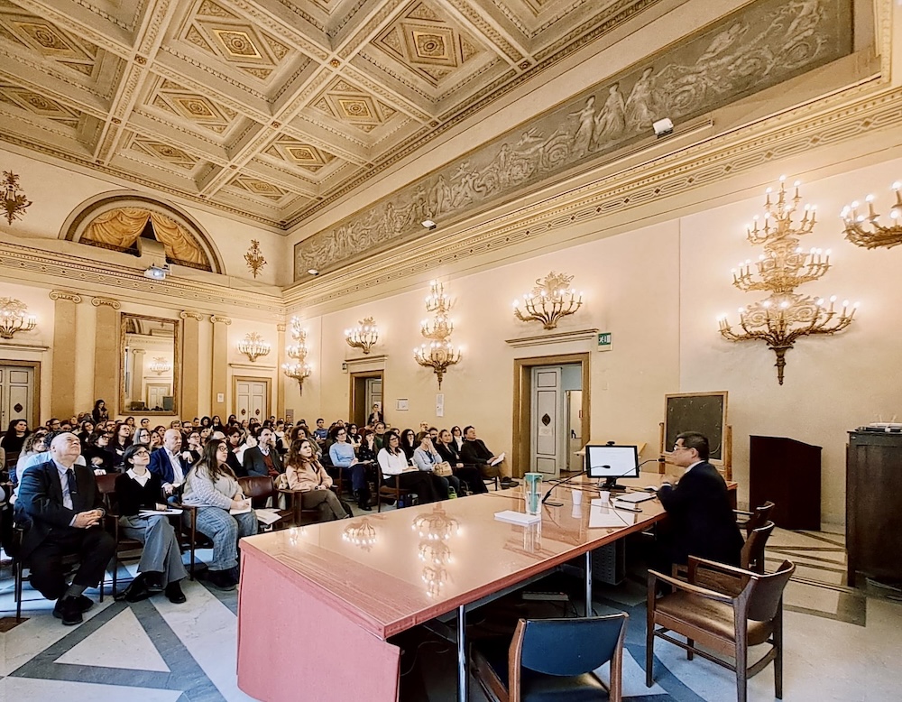 A spacious, ornate room with a high ceiling, elaborate moldings, and wall sconces. An audience sits attentively facing a long table where a speaker is presenting near a computer. The room has a classical style with a mural above the walls and large chandeliers.