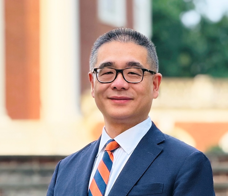 Portrait of a person in a blue suit and orange striped tie, standing confidently in front of a blurred historic building.