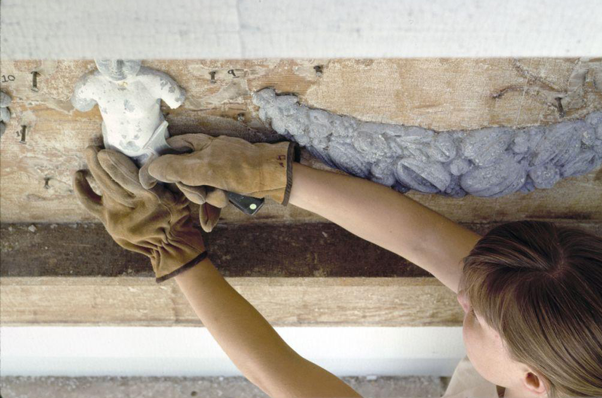 An individual wearing gloves works on cleaning a frieze. The frieze features a figure and ornate patterns.