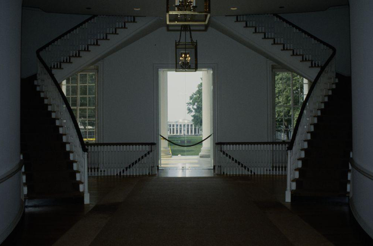 A grand entrance hall in the University of Virginia Rotunda with dual staircases curving upwards on each side. Large windows flank a central open doorway, revealing a view of the UVA Lawn with a tree, greenery, and a white columned building. Elegant chandeliers hang from the ceiling.