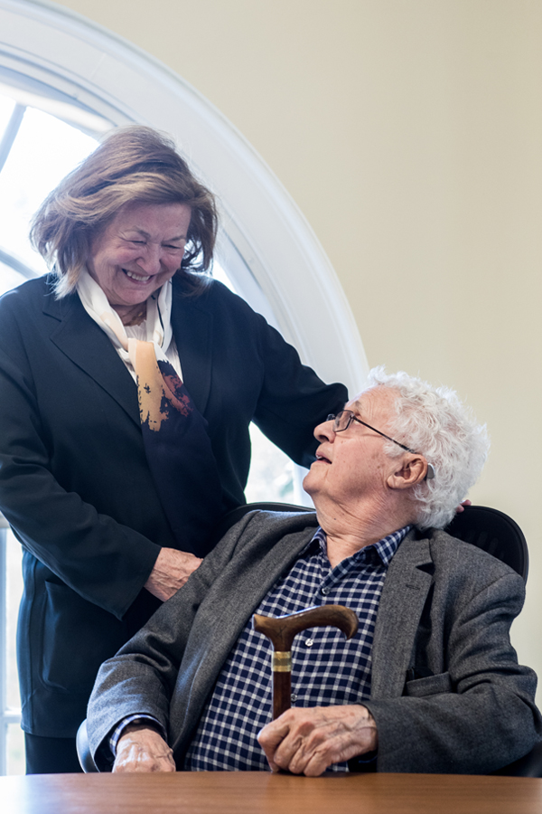 Two people are in front of a arched window. A woman, standing and wearing a black jacket and white shirt, beams down at a seated man with glasses and a checkered shirt and gray blazer. He smiles back up at her.