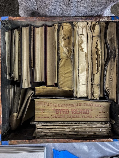 A collection of worn and aged books is stacked vertically and horizontally in a wooden crate. One visible book cover reads "The Haxall-Crenshaw Company" followed by "Byrd Island Patent Family Flour." The books show signs of wear, with some pages and covers exhibiting creases and discoloration.