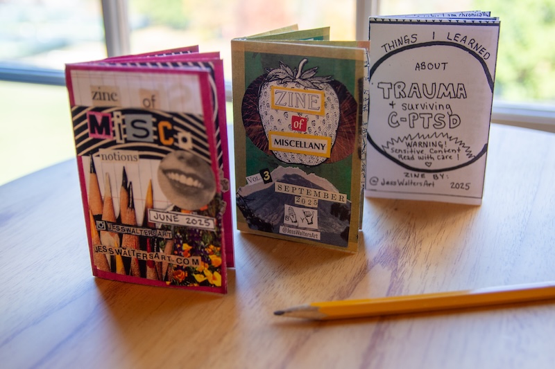 Three zines titled "Miscellany of Emotions", "Zine of Miscellany", and "Things I Learned About Surviving C-PTSD" alongside a pencil, displayed on a wooden table near a window.