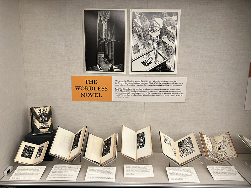 The image shows a display titled "The Wordless Novel" in an exhibition. At the top, there are two large black-and-white woodcut-style illustrations. Below these, a caption explains the history and concept of wordless novels, mentioning artists like Lynd Ward, who popularized this genre. Several open books are displayed beneath the caption, each containing black-and-white illustrations, emphasizing the visual storytelling aspect of the genre. One of the books on the left has the title "Gods' Man" by Lynd Ward, and each of the books is propped open on individual stands to reveal their intricate artwork.