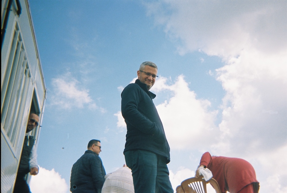 A person stands outdoors looking downward, with hands in pockets, wearing glasses and a dark jacket. Two others are nearby; one is seated, arranging items on a plastic chair. A truck with a visible open door is in the background. The sky is partly cloudy.