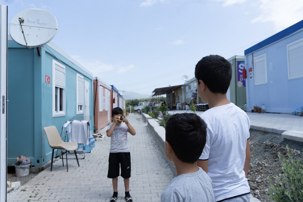 A group of young people outdoors on a paved path lined with temporary housing units. One individual is using a small camera. A Turkish flag is visible on one of the units. Satellite dishes and a chair are nearby. The sky is partly cloudy.