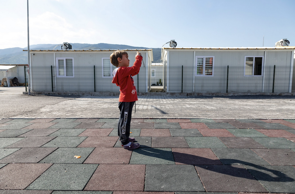 A child stands outside, holding a camera. In the background, there are two living containers and a fence, with a mountain range visible under a clear sky.