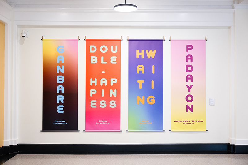 Four colorful banners hang against a white wall. They read: Ganbare, Japanese, to persevere; Double Happiness, Chinese, joy and unity; Hwaiting, Korean, you got this!; Padayon, Visayan dialect / Philippines, to carry on