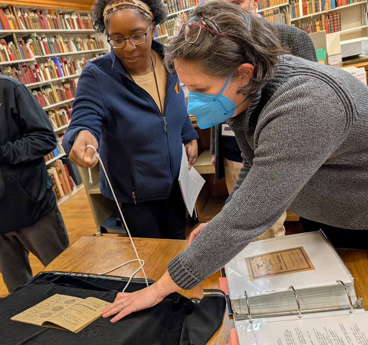 Two Library staffers carefully display a small, discolored book. A binder holding more historic items is open on the table.