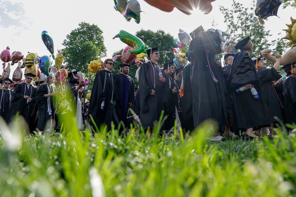 A group of graduates in caps and gowns walk across a grassy area, surrounded by colorful balloons. The sun is shining through the trees in the background.