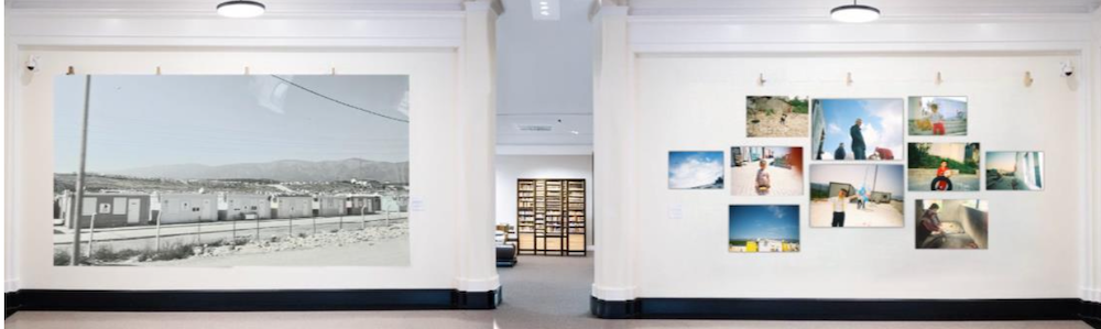 This image shows a gallery exhibit with photographs displayed on walls. On the left, a large black and white photo of a housing area is displayed. On the right, multiple smaller color photographs are arranged in two rows.