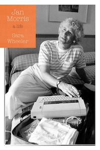 A person sitting on a bed wearing a striped shirt and looking upward, next to a typewriter. The cover includes the text "Jan Morris a life" by Sara Wheeler.