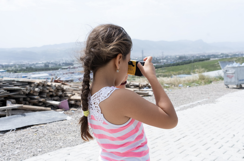 A person with long hair in a striped tank top stands on a paved area, taking a photo with a camera. In the background, there are piles of wooden planks and a view of a cityscape with mountains.