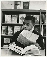 Young man sitting at a table in front of a bookshelf with brochure entitled: “Observe Negro History Week with a book...”