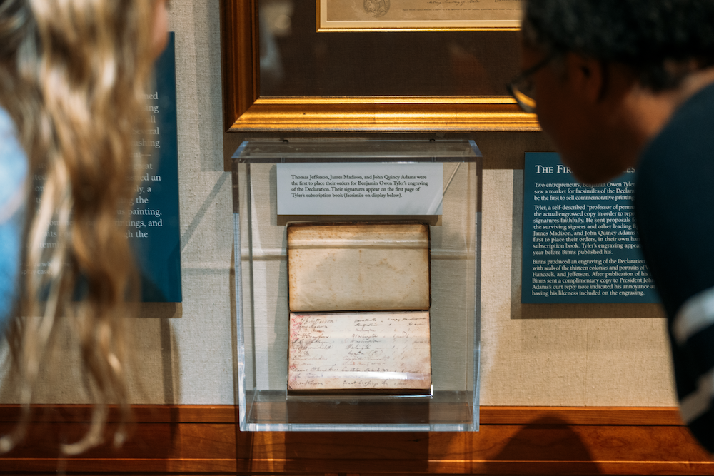 Two people are examining a historical document displayed in a glass case at a museum. The document is related to Thomas Jefferson, James Madison, and Gilbert du Motier, Marquis de Lafayette. Labels and descriptions are mounted on the wall, providing context about the exhibits.