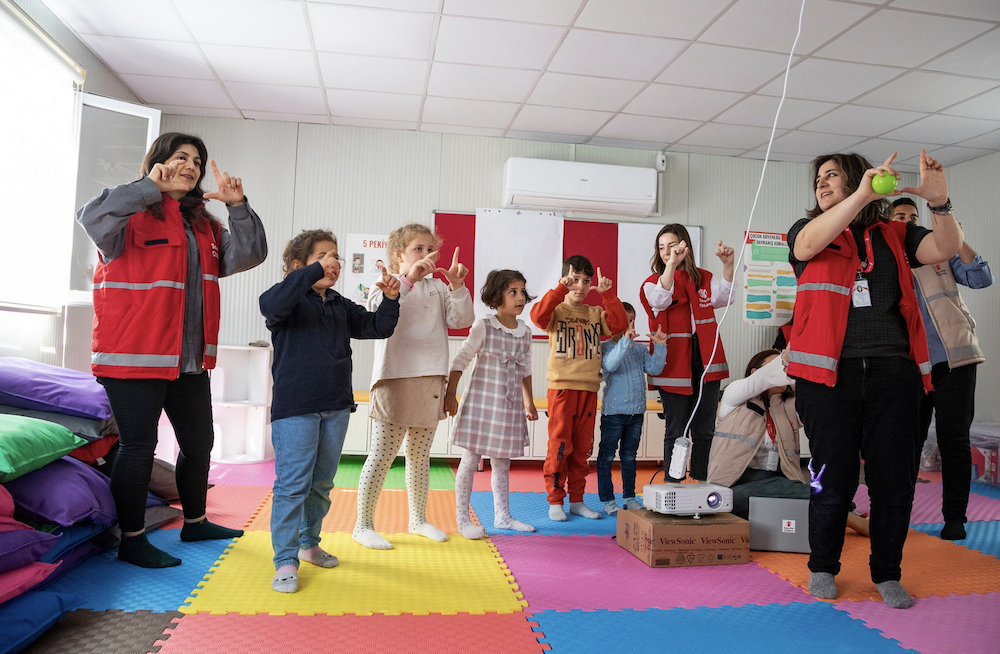 A group of people and children stand on a colorful foam mat floor, engaging in playful activities. Several people are wearing red vests with "Save the Children" written on them. The room is well-lit and includes various cushions and a ceiling-mounted air conditioner. A few individuals are making hand gestures for the children to mimic.