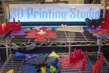 A display of various 3D printed objects on a wire rack. Behind is a lit 3D printed sign that reads "3D Printing Studio"