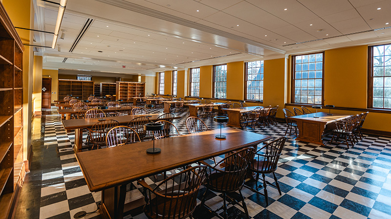 Long view looking down a reading room with black and white checkered floors and wooden chairs accompanying long wooden tables with 2 lamps on each table.