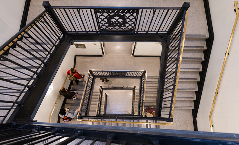 View in the interior of a building looking down several floors from the center of a stairway with landings at right angles to the steps.