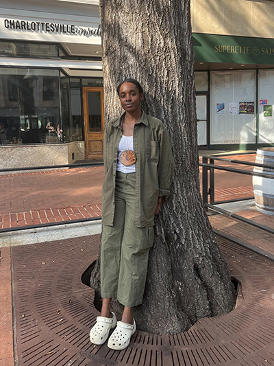 Person standing by a tree on a pedestrian street, wearing a long green coat over a white t-shirt with a sun design, and white shoes. Shops are visible in the background.
