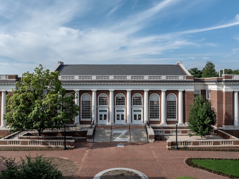Exterior view of Shannon Library at the University of Virginia on a sunny day, featuring a grand staircase leading up to multiple entry doors flanked by large columns, set against a clear blue sky.